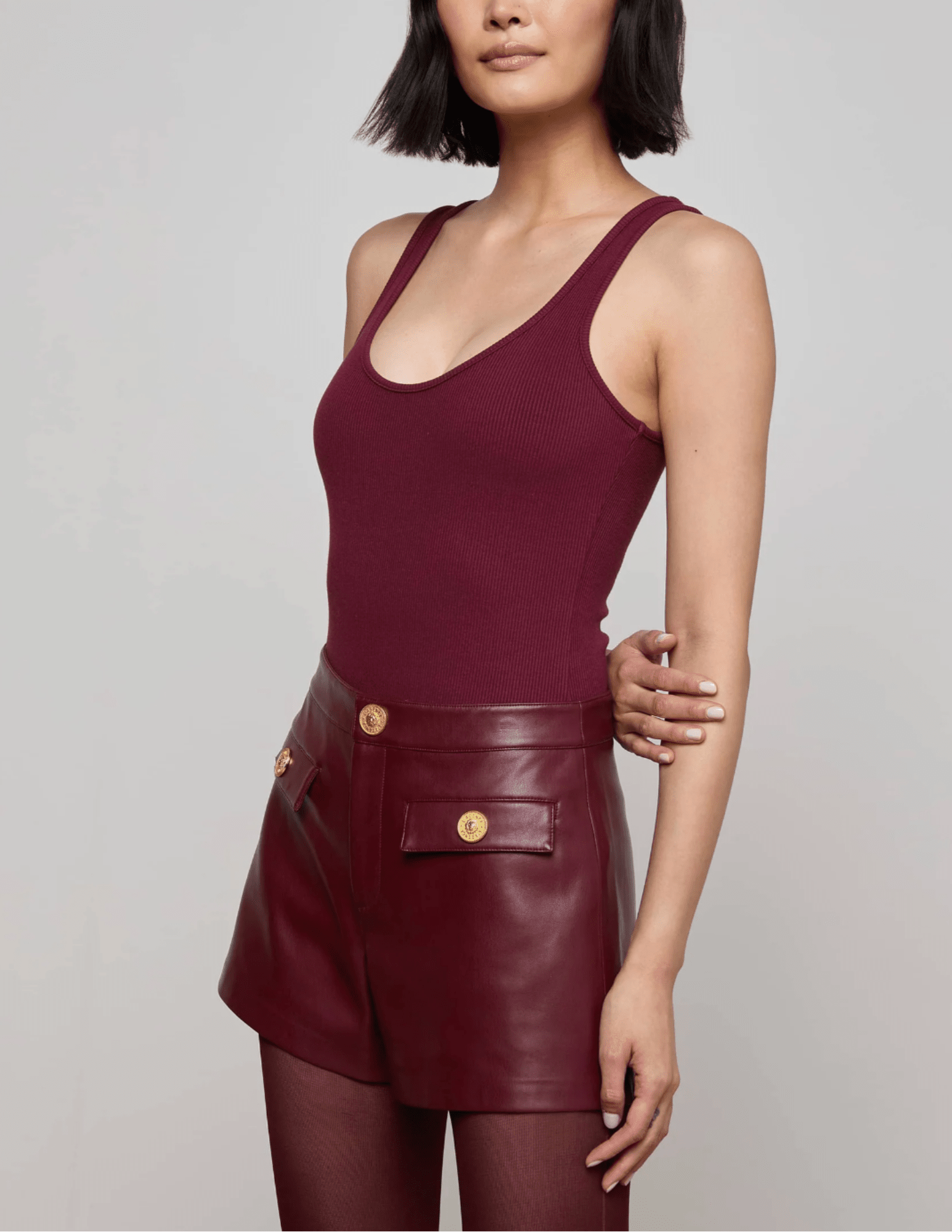 Side view of model wearing L’AGENCE Nica Scoop Neck Bodysuit in Dark Port with leather shorts.
